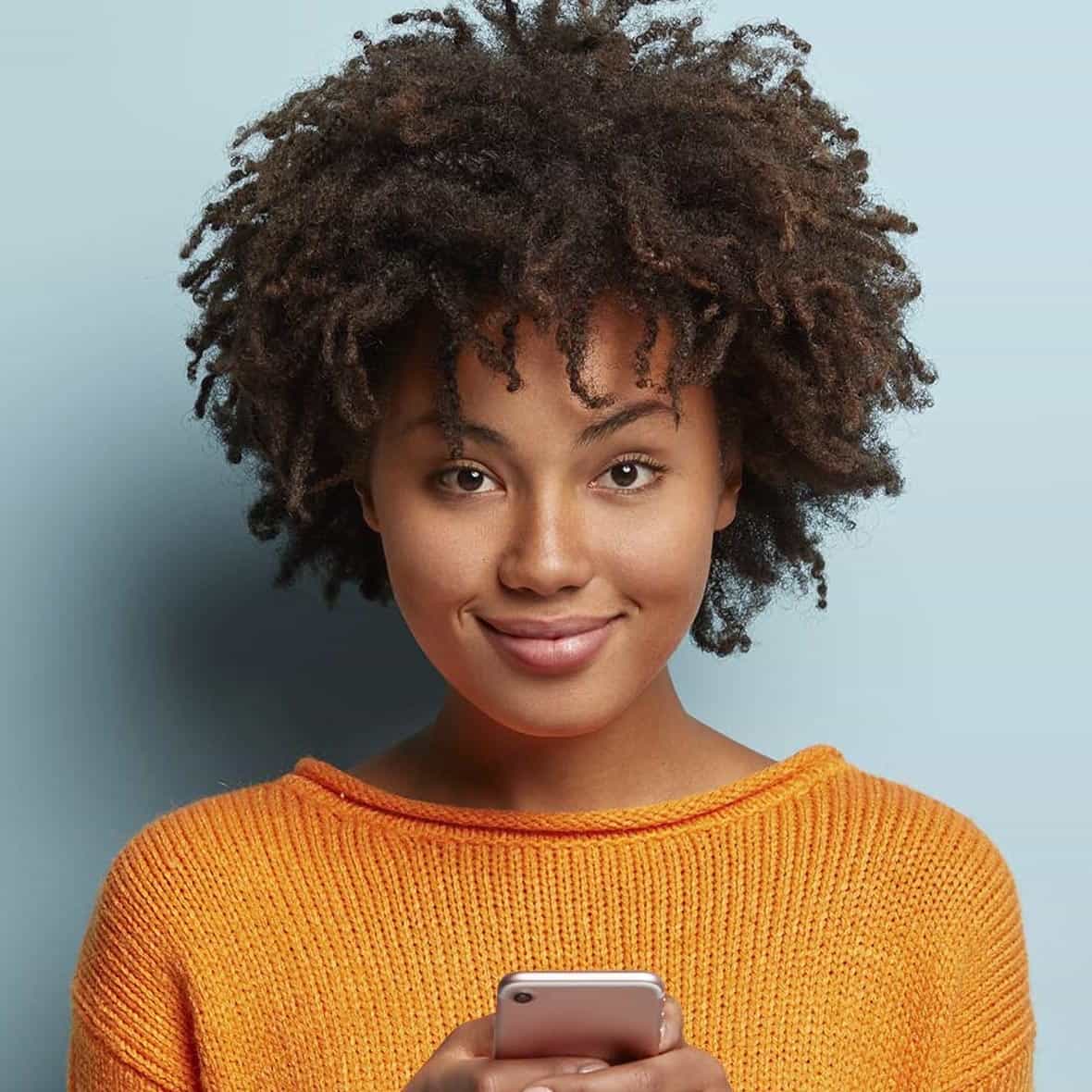 Chica afroamericana con el pelo largo y rizado con un jersey naranja sosteniendo un móvil