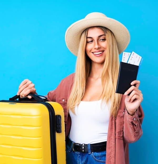 Mujer rubia de pelo largo con un sombrero sosteniendo billetes de avión y una maleta amarilla